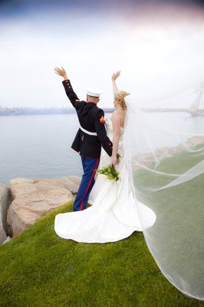 Newlyweds, Marine in uniform and bride in white dress, raise arms on a cliff by the water.
