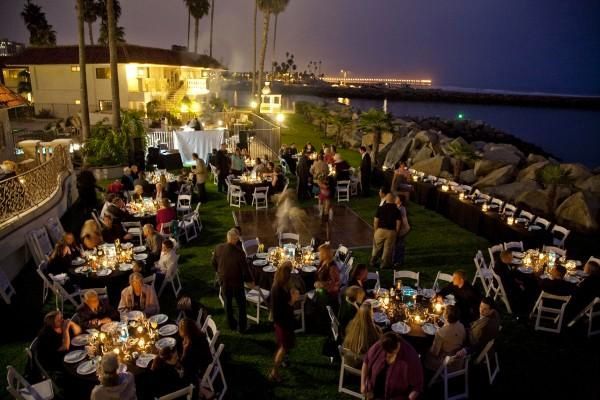 Outdoor evening event with people at tables, overlooking water. Dim lighting, tables with candles.