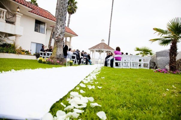Wedding ceremony on green grass, white aisle runner, guests seated, gazebo in the background.