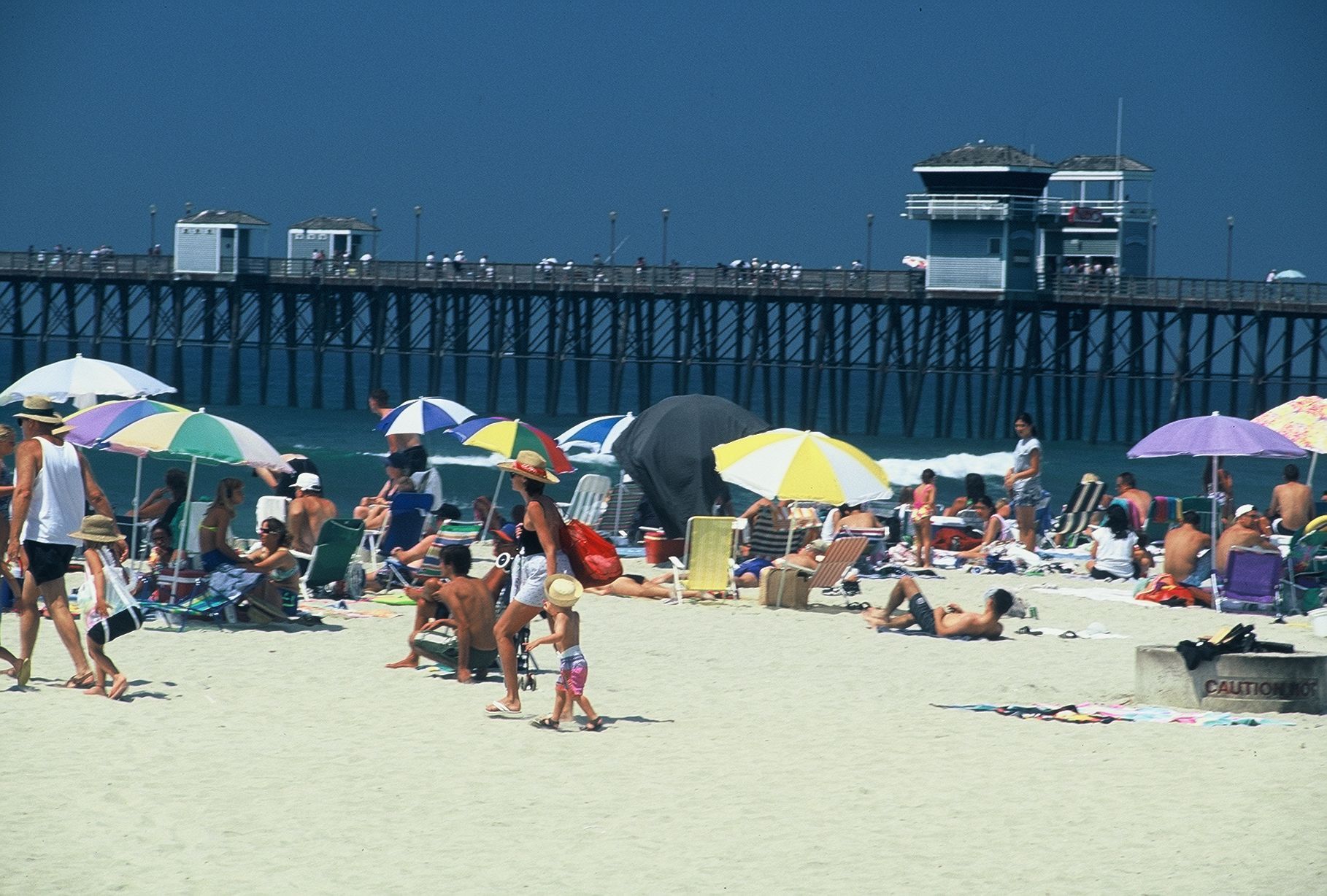 Beach scene with pier in background, people under umbrellas, relaxing on the sand.