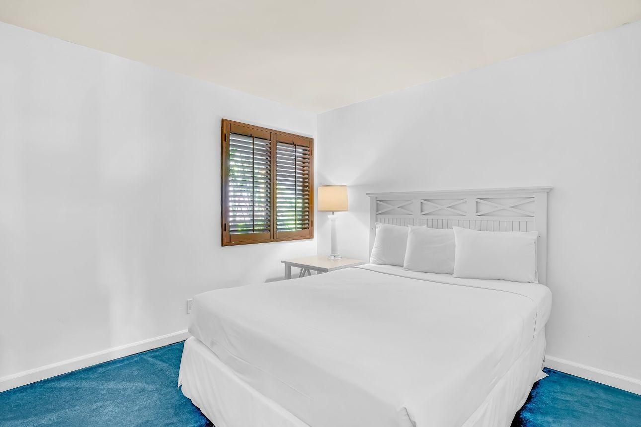 Bedroom with white walls, bed, window with wood shutters, blue carpet.