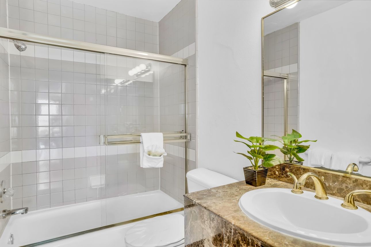 Bathroom with a white tub, shower, and sink, marble countertop, gold fixtures, and small potted plant.