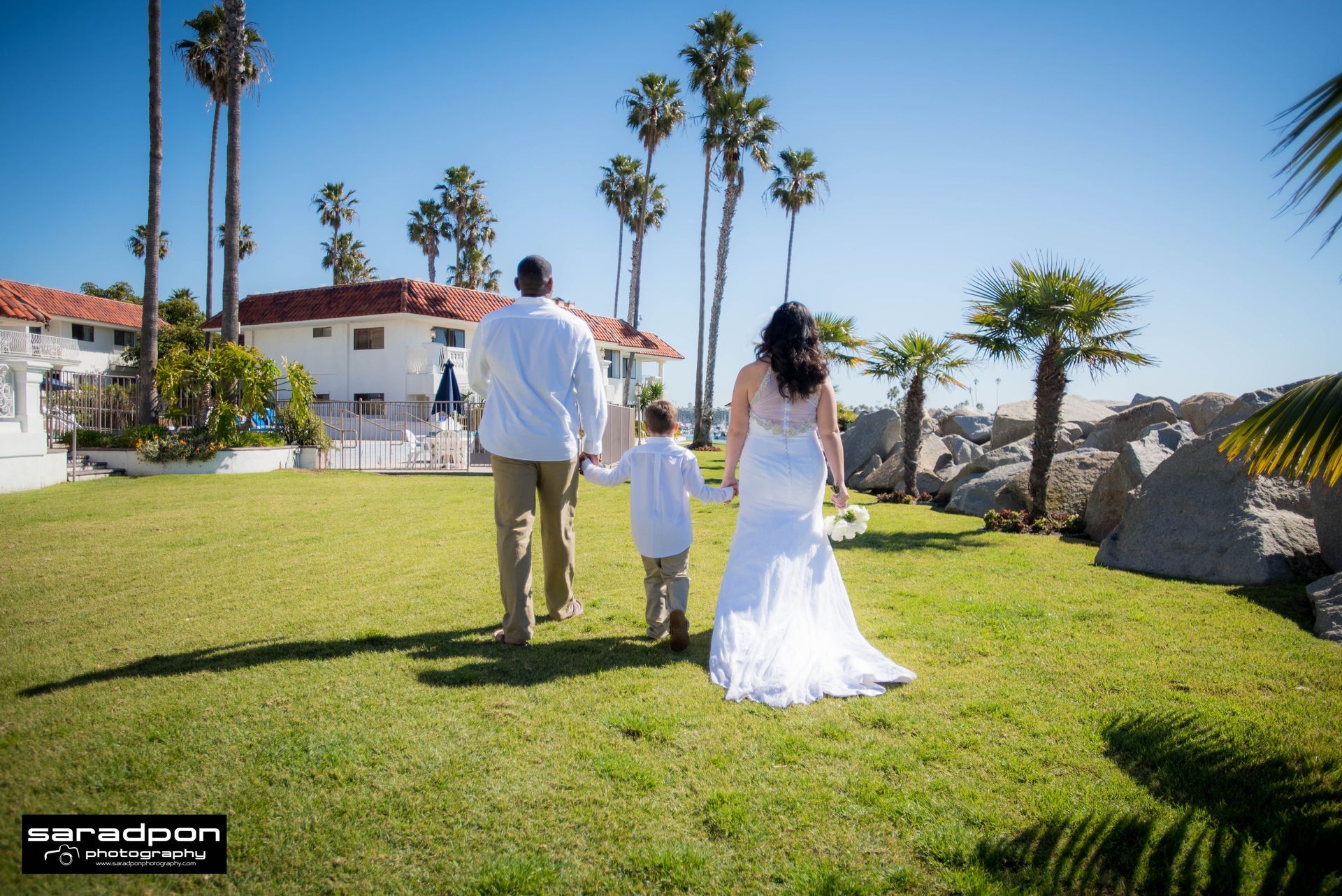 Family walks on grass toward the ocean: man, woman in wedding dress, child. Sunny day, palm trees, white building.