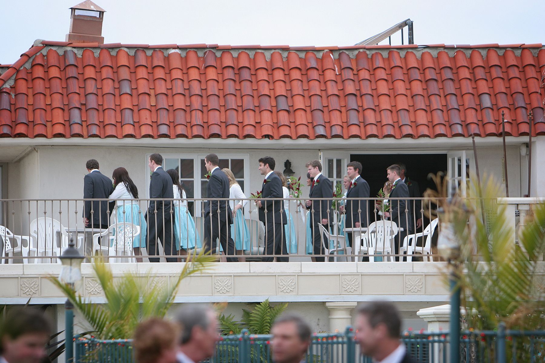 Wedding party on a balcony; suits and turquoise dresses, white building, red tile roof, outdoor setting.