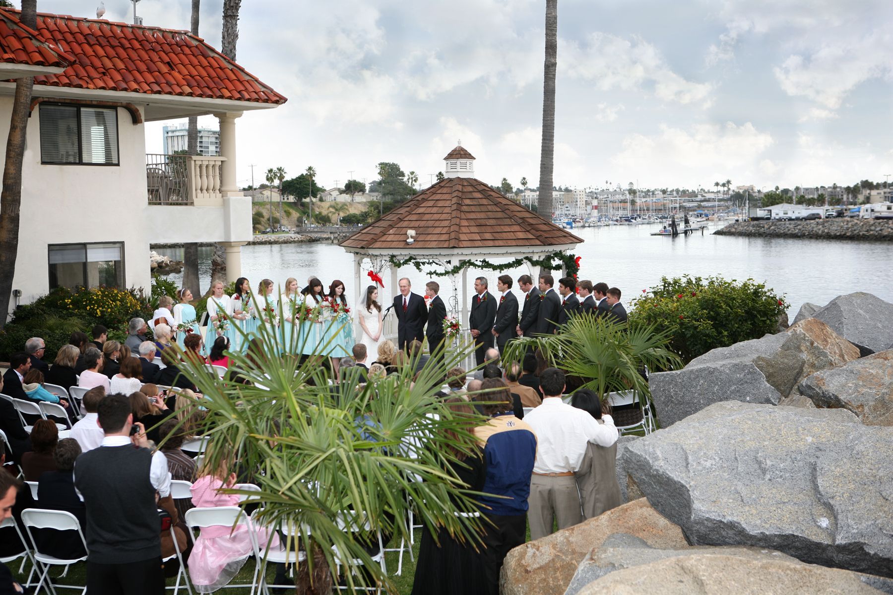 Wedding ceremony outdoors by a harbor. Bride and groom near small gazebo; guests seated.
