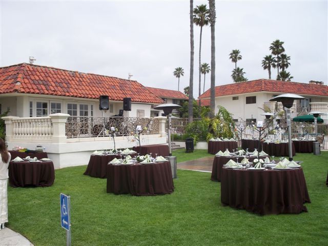 Outdoor event with tables covered in brown linens, set up on grass, by a building with a red-tiled roof.