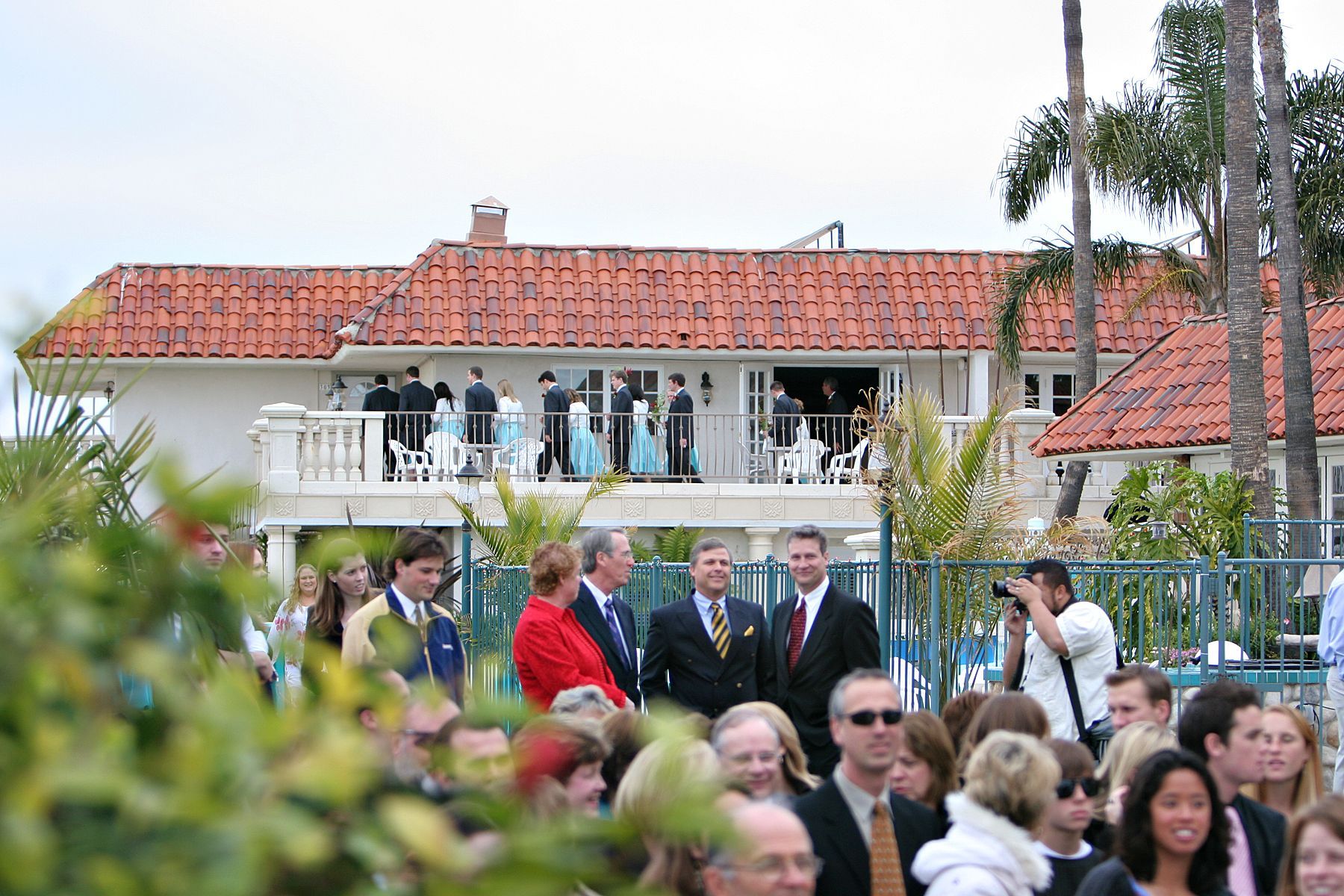 People gathered outside a white house with a red-tiled roof; some are on a balcony.