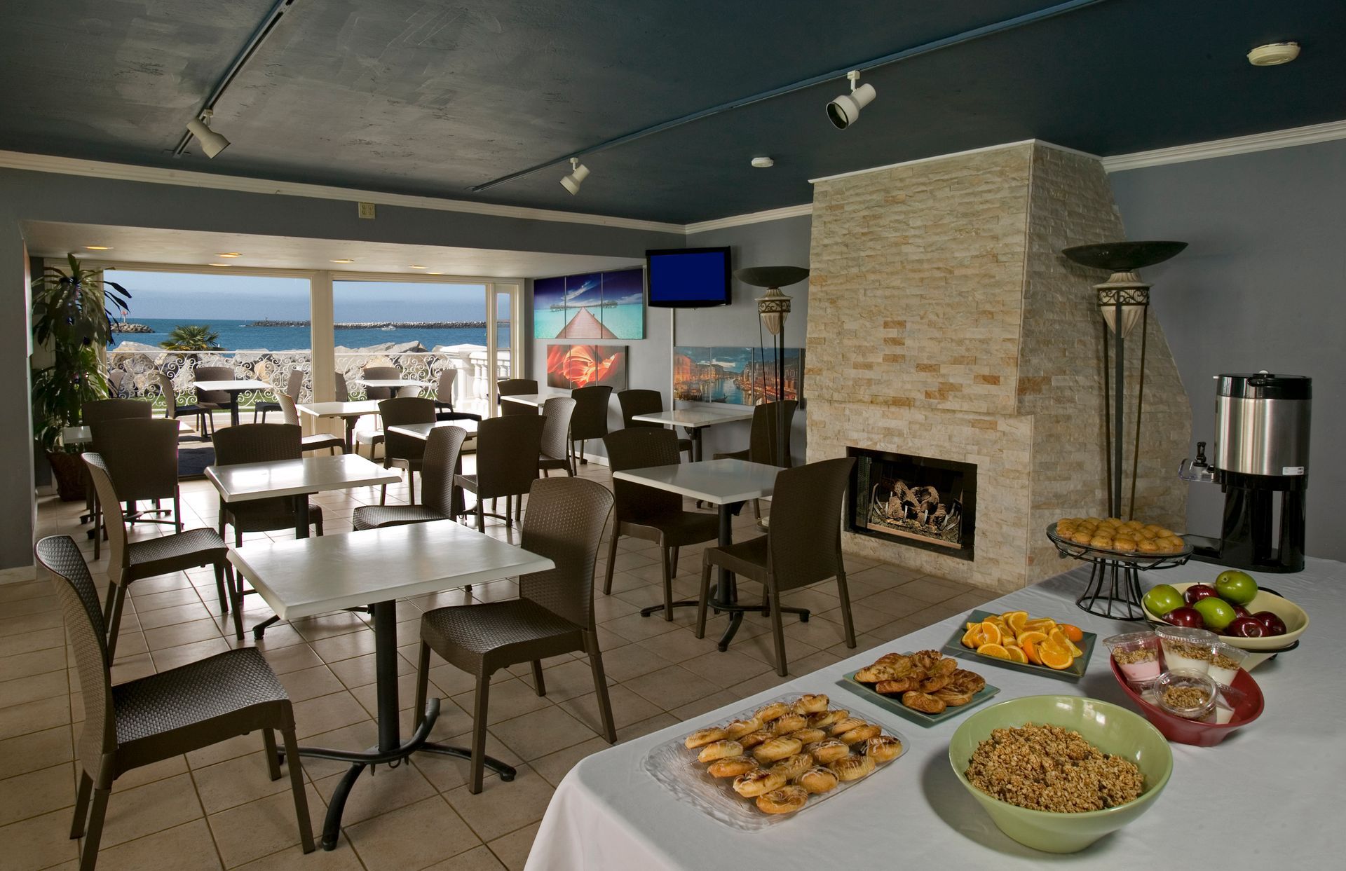 Hotel breakfast area with food buffet, tables, chairs, fireplace, and ocean view.
