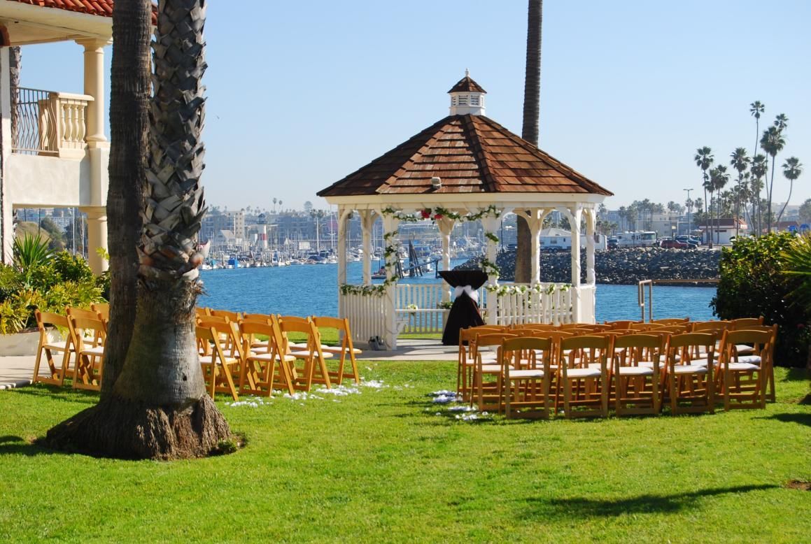 Wedding ceremony setup: gazebo, wooden chairs on green lawn overlooking harbor, sunny day.
