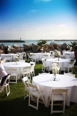 Outdoor wedding reception with white tables and chairs on grass, ocean view.