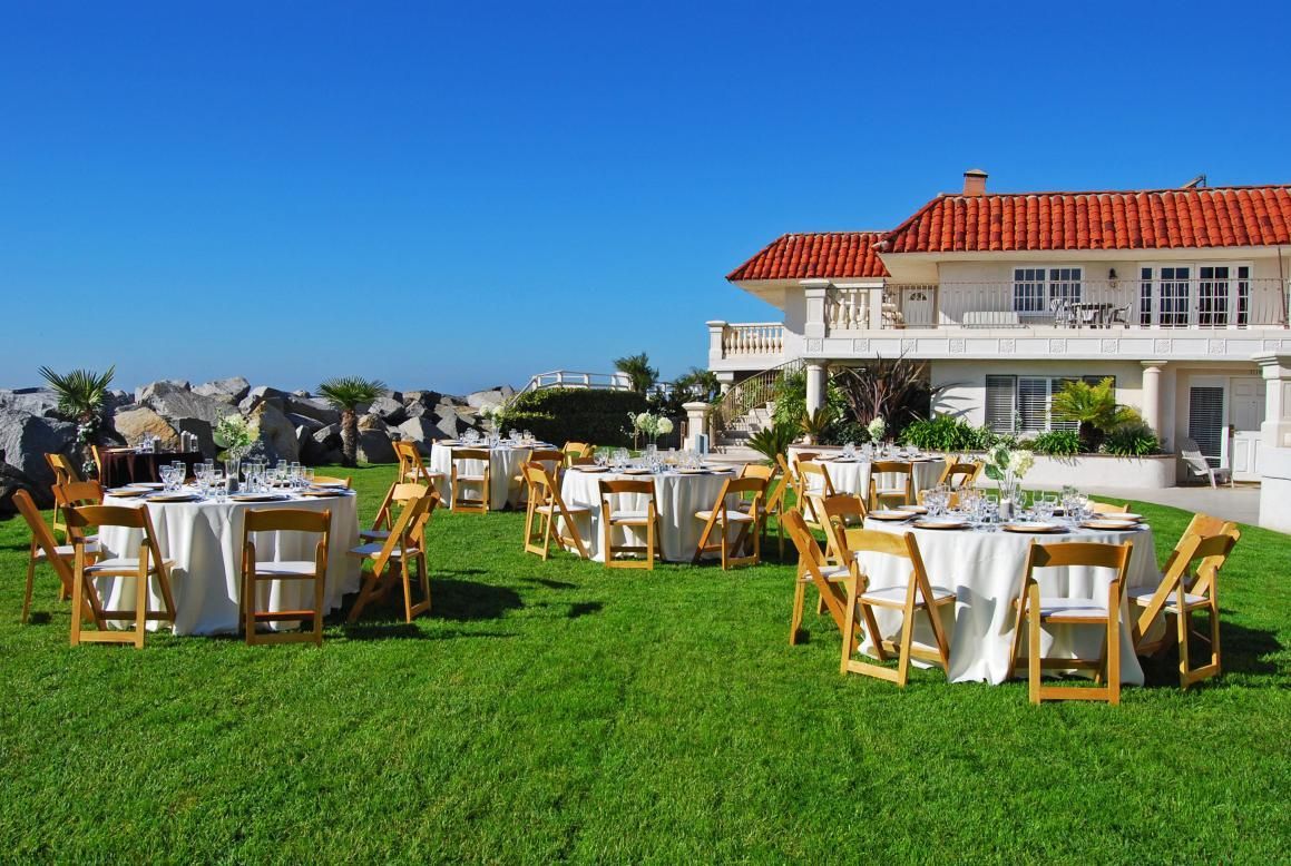 Outdoor event setup with round tables on grass, a white building, and blue sky.