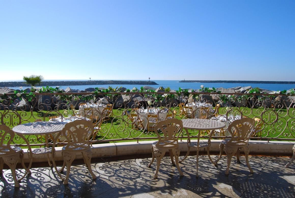 Outdoor cafe with ocean view, gold chairs, tables, green grass, blue sky.
