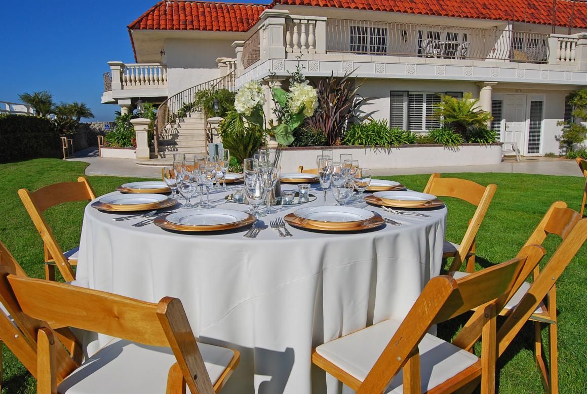 A round table set for a meal in a grassy yard, with a large white house in the background.