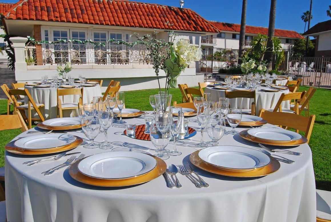 Round tables set for an outdoor event, featuring white linens, gold chargers, and floral centerpieces.