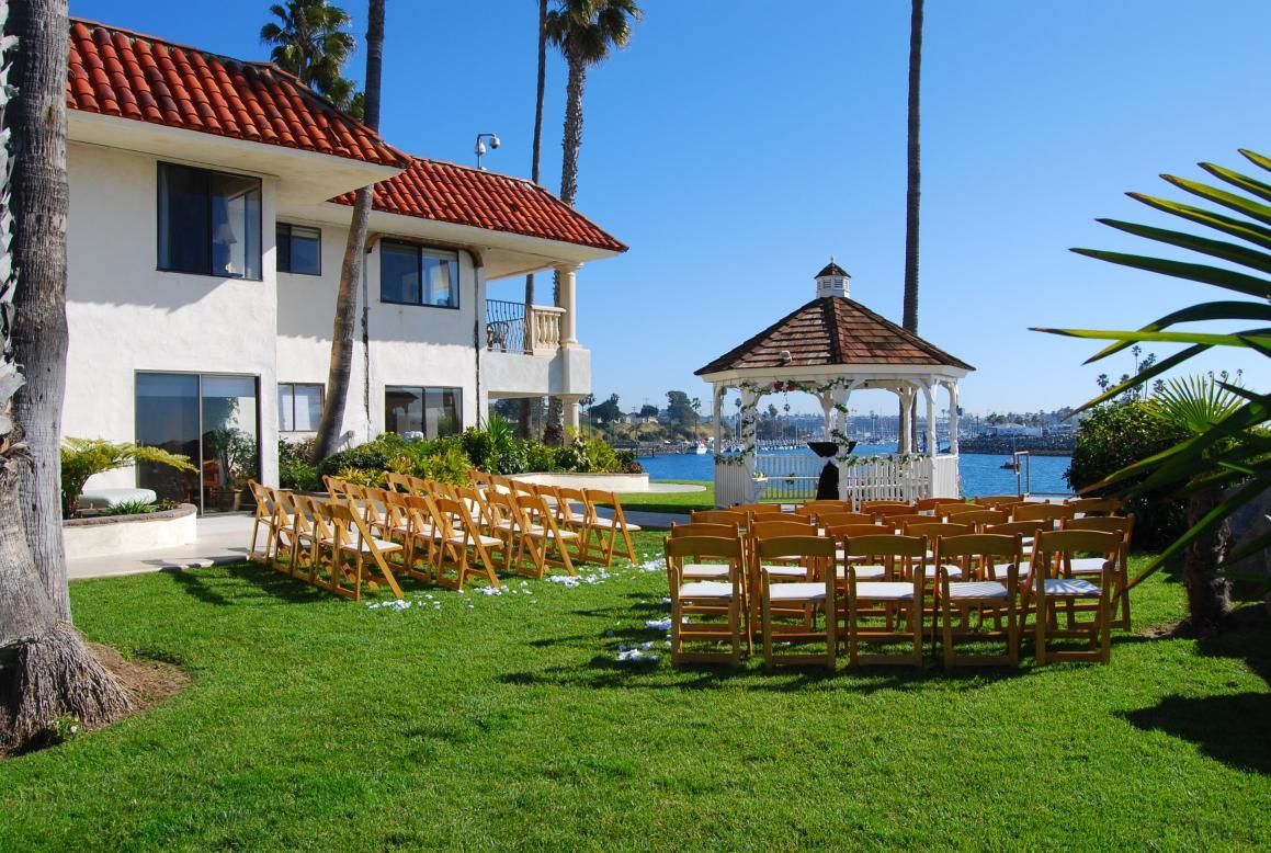 Wedding ceremony setup on a grassy lawn with a gazebo, facing the ocean, with a white building.