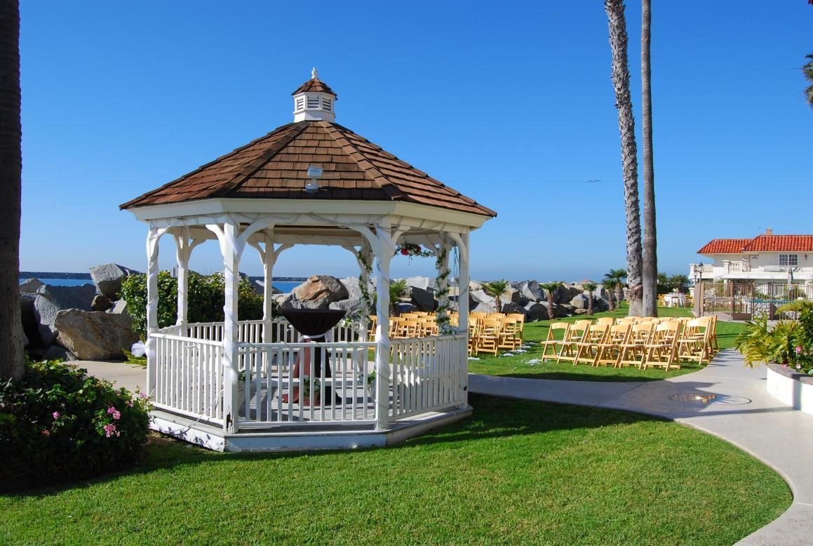 White gazebo on green lawn, wedding chairs set up. Ocean and sunny blue sky in background.