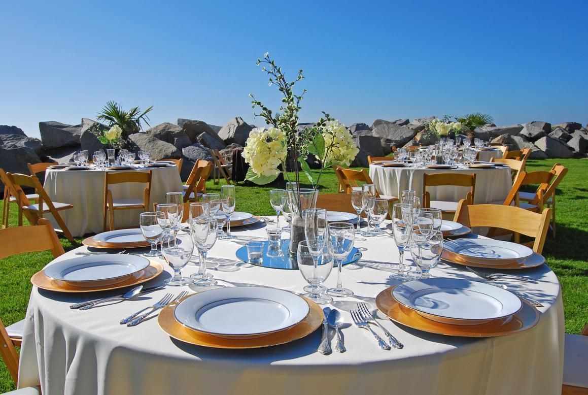 Wedding reception table set outdoors, white linens, gold chargers, clear glasses, floral centerpiece, blue sky.