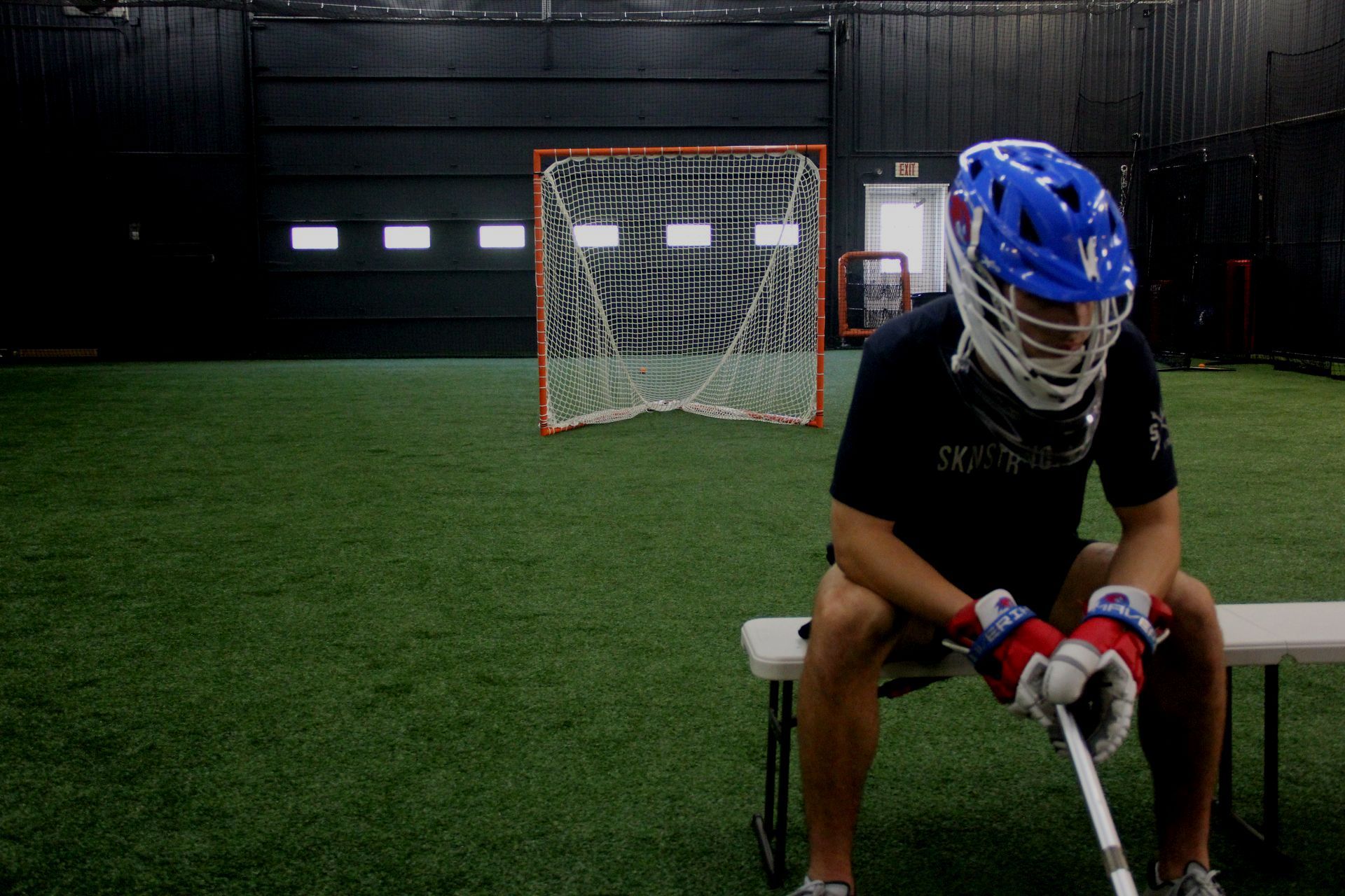 A lacrosse player is sitting on a bench holding a stick.