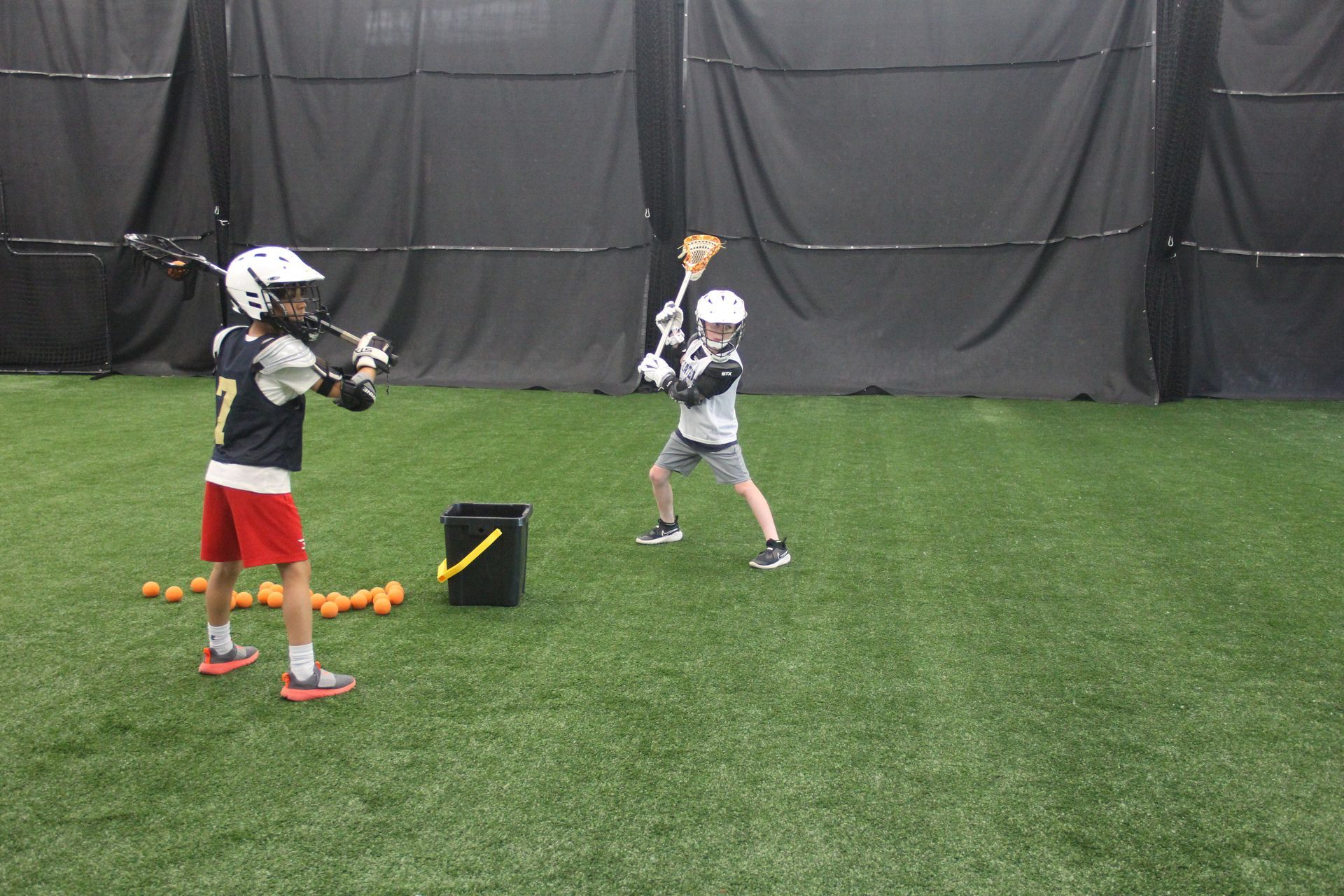 Two young boys are playing lacrosse in a cage.