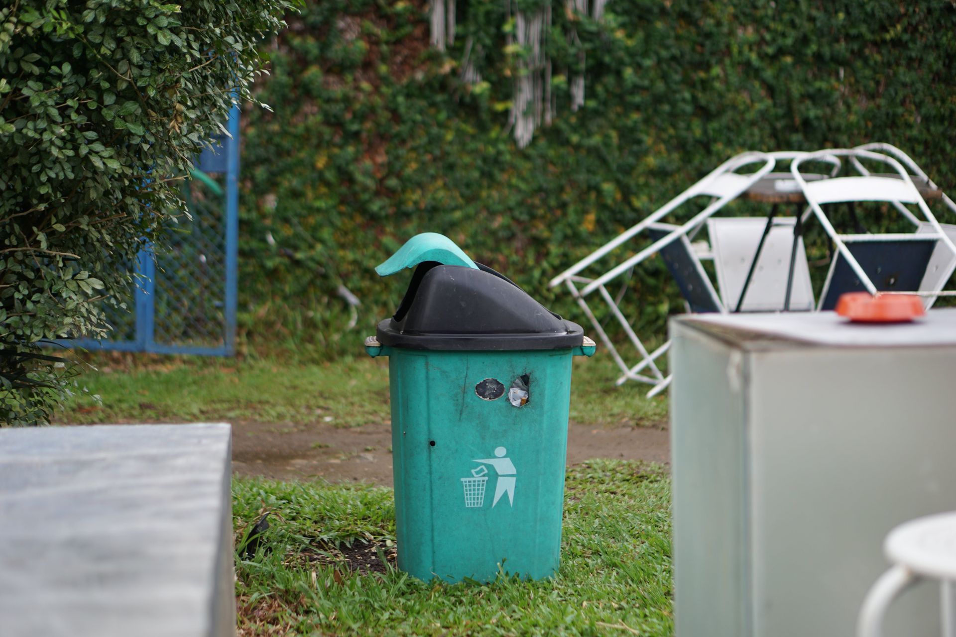 a green trash can is sitting in the grass next to a broken chair .