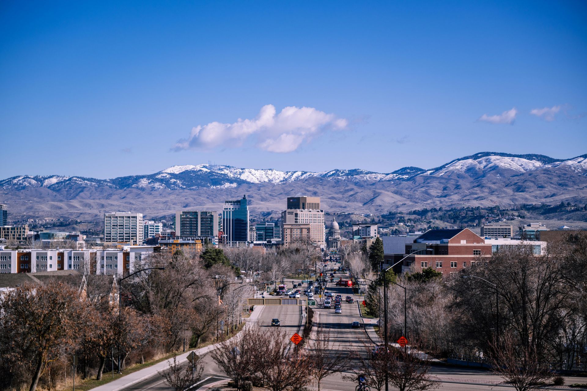City skyline with modern buildings, a bustling street, and snow-capped mountains under a clear blue sky.