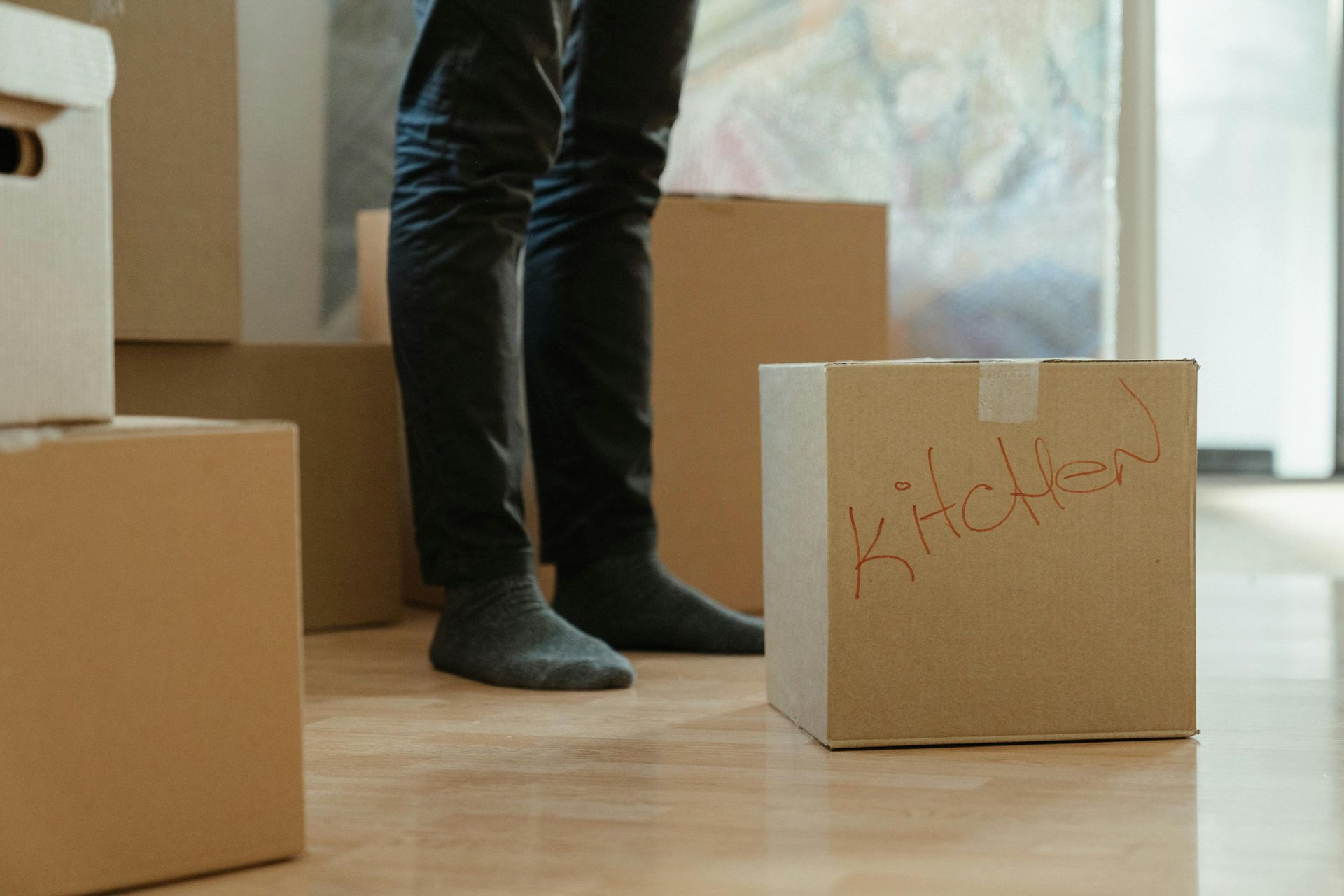A person in dark pants and gray socks stands among cardboard boxes, one labeled 