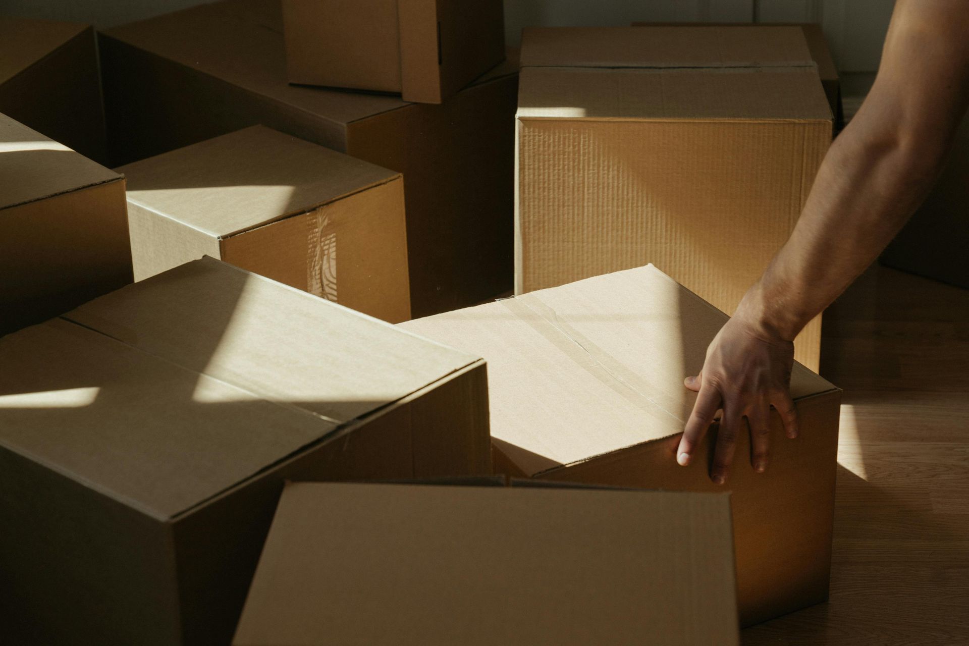 A hand places a brown cardboard box onto a stack of similar boxes on a wooden floor, lit by natural sunlight.