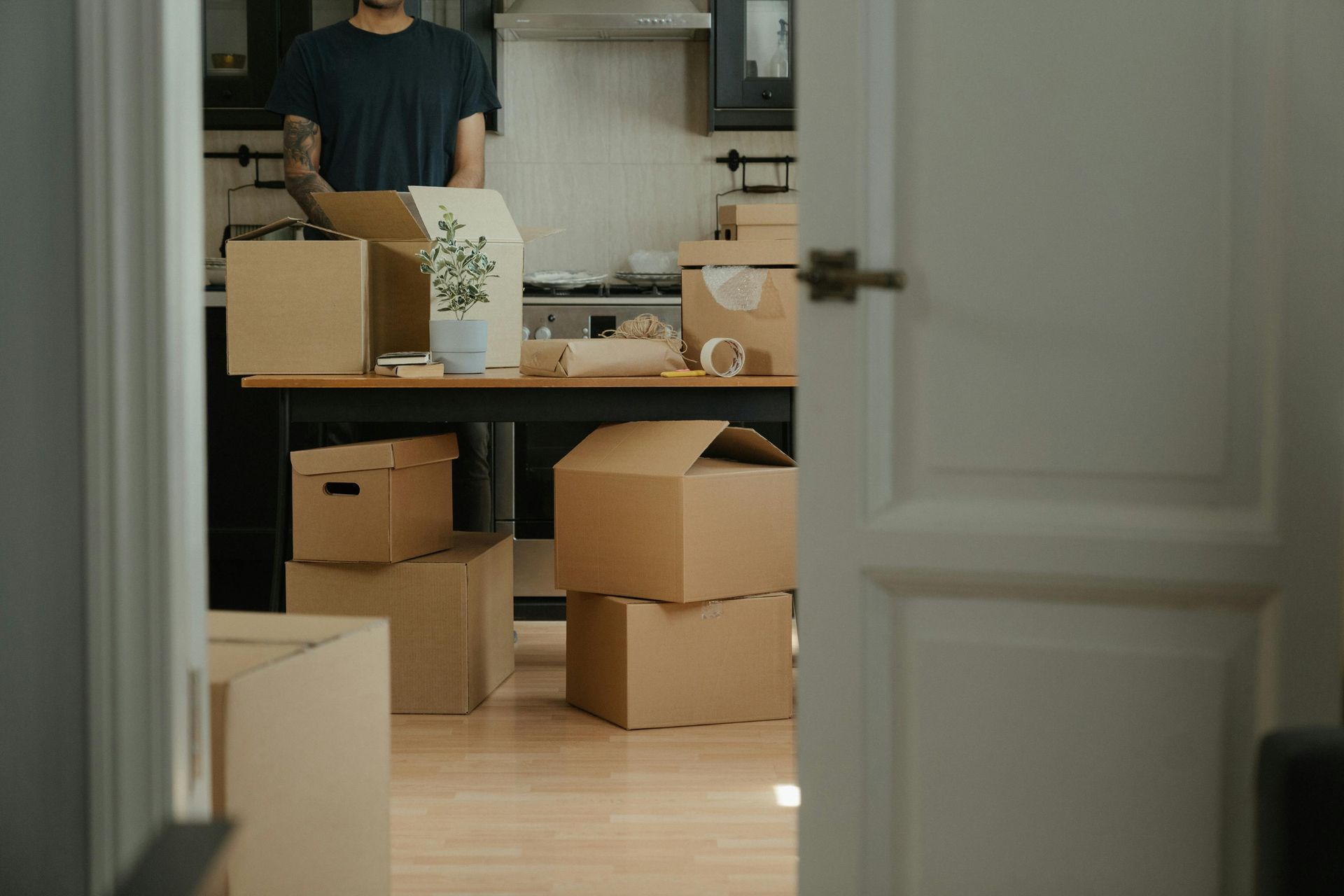 A person packs cardboard boxes on a table in a kitchen filled with more boxes, viewed through an open doorway.