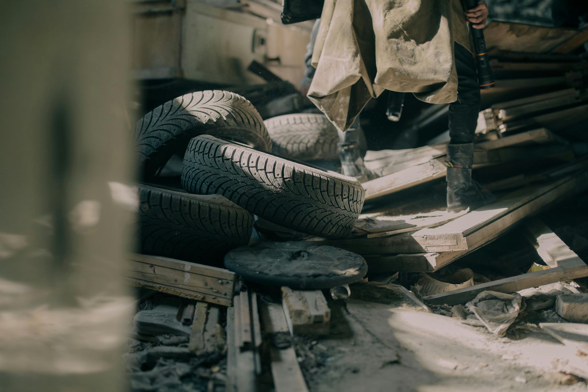 A person in a beige coat and dark boots walks through debris, including discarded tires and wooden planks.