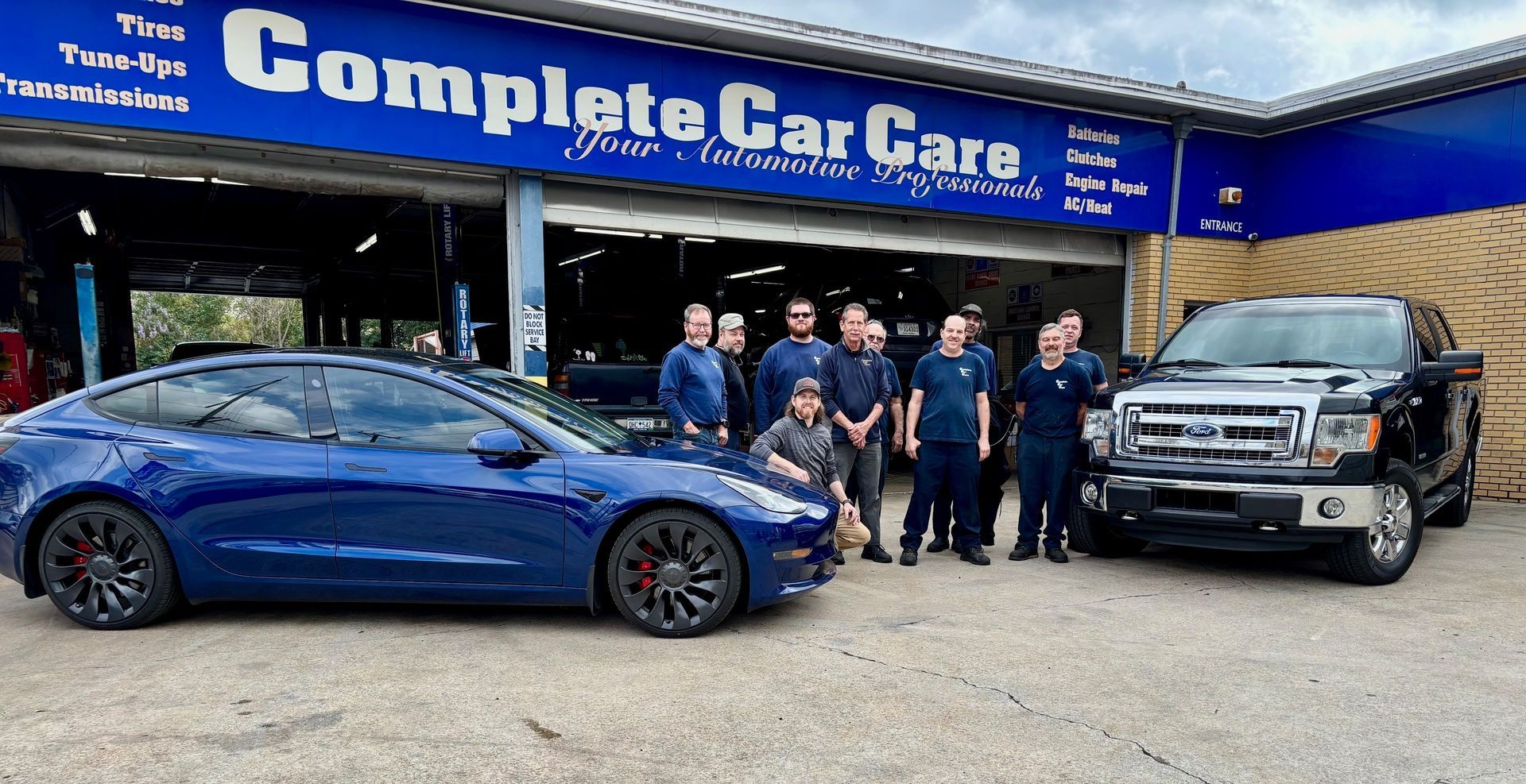 A team of automotive professionals posing in front of a Complete Car Care auto repair shop, with a blue Tesla Model 3 and a black Ford F-150 parked in front of the garage.