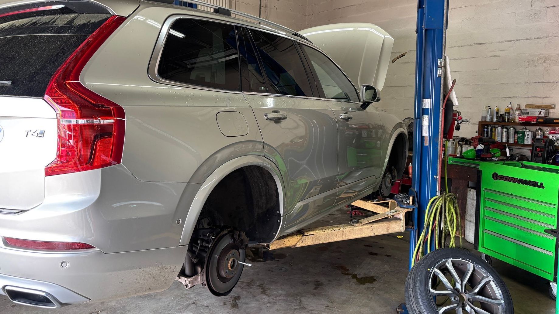 A silver Volvo XC90 SUV lifted on a hydraulic car lift in an auto repair shop, with its front hood open and wheels removed for maintenance. A green tool chest and various automotive tools are visible in the background