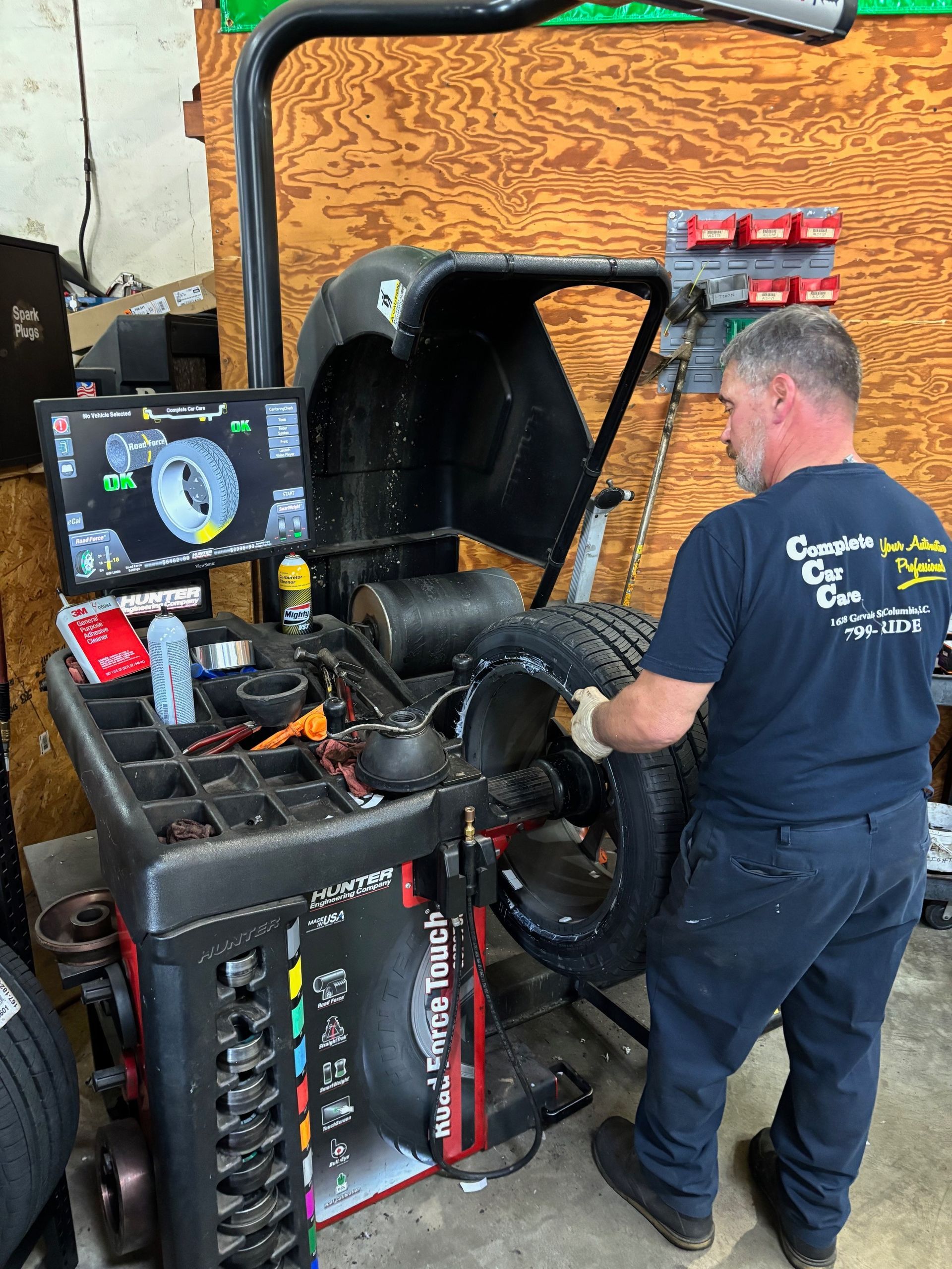 An auto technician wearing a 'Complete Car Care' shirt balances a tire using a Hunter Road Force Touch wheel balancer in a workshop. The machine's screen displays alignment and balance readings, while various tools and supplies are arranged nearby.