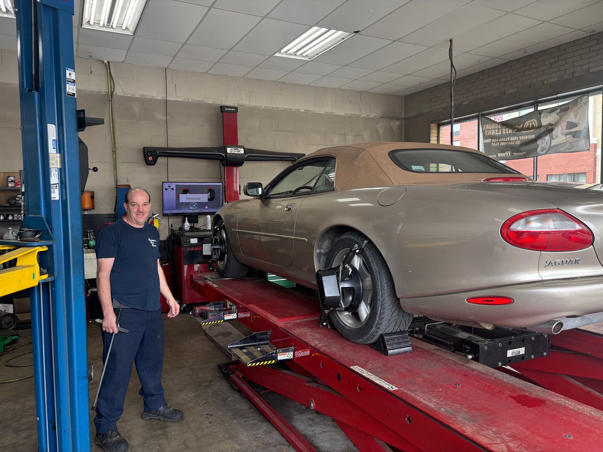 An auto technician stands next to a beige Jaguar convertible on a red hydraulic lift in a workshop, preparing for a wheel alignment. The car's wheels are fitted with alignment sensors, and a digital screen displays measurement data.