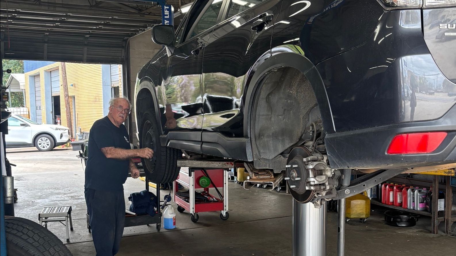 A mechanic working on a dark-colored Subaru SUV lifted on a hydraulic post lift inside an auto repair shop. The vehicle's wheels are removed, and hubcaps and tools are scattered on the concrete floor.
