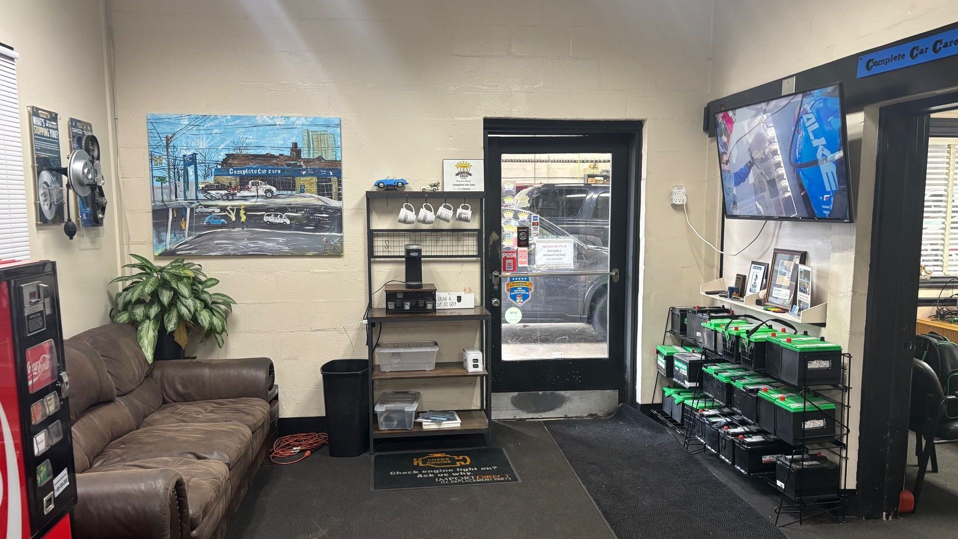 An auto repair shop waiting area with a brown leather couch, a red vending machine, and a wall-mounted TV displaying a security camera feed. A shelf with coffee supplies is near the door, and car batteries are neatly stacked on racks. A large painting of the shop hangs on the wall.