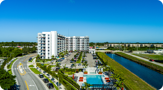 Aerial view of a white apartment complex beside a canal, road, parking lot, and pool under a clear blue sky
