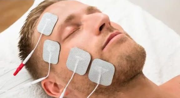 Hombre con electrodos en la cara, recibiendo tratamiento de estimulación facial, tumbado sobre una superficie blanca.