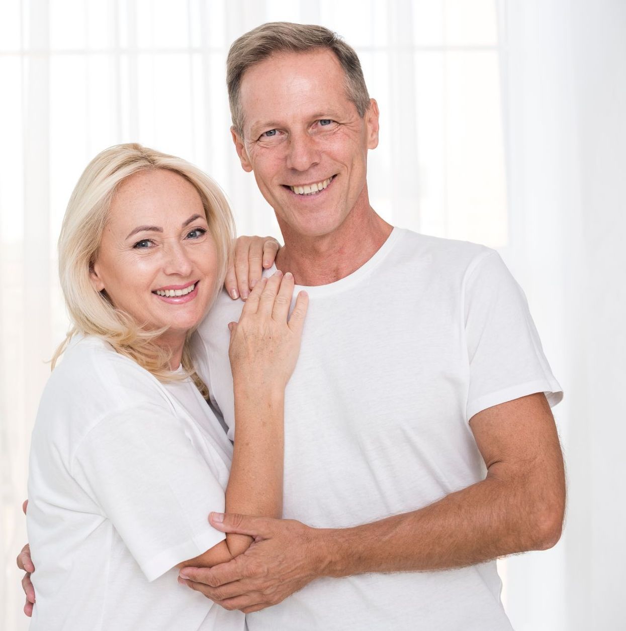 Pareja sonriente, abrazada frente a un fondo blanco, vistiendo camisetas blancas.