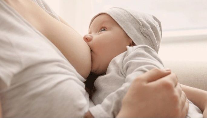 Bebé mamando de una persona, en interiores. El bebé lleva un gorro y mira hacia un lado.