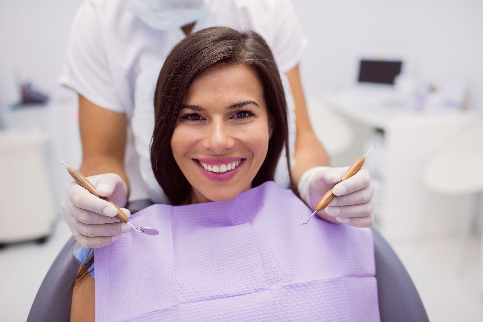 Una mujer sonríe sentada en el sillón del dentista durante una revisión. El dentista sostiene los instrumentos; fondo blanco.
