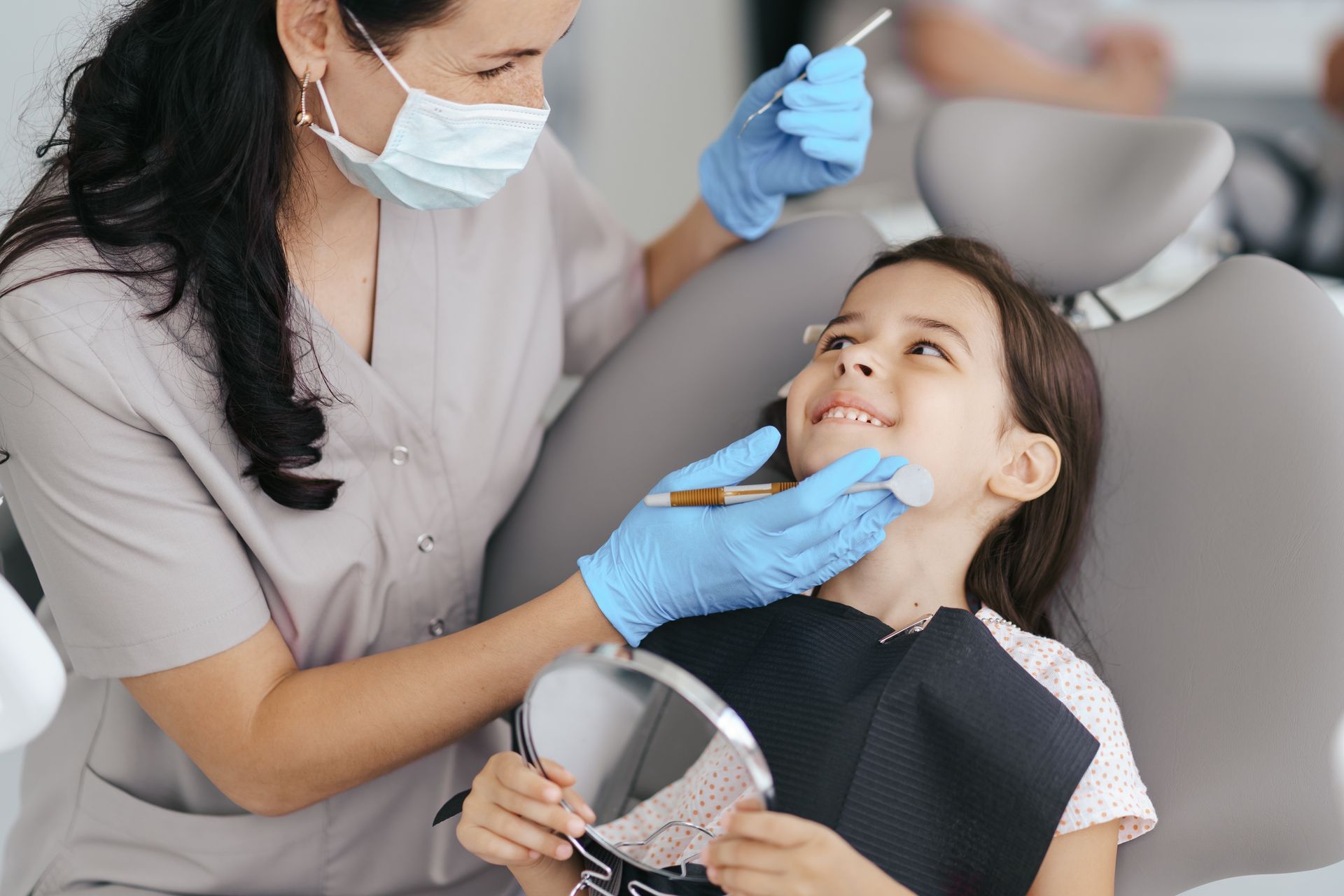Un dentista examina los dientes de un niño sentado en un sillón dental; ambos sonríen; hay instrumental dental y un espejo presentes.