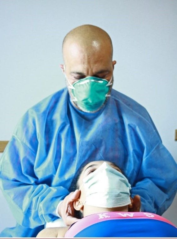 Médico con bata azul y mascarilla atendiendo a una persona acostada, que también lleva mascarilla.