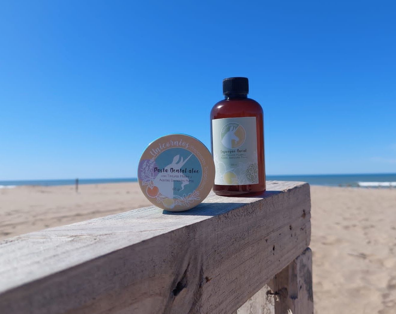 Frascos de jabón y loción sobre una superficie de madera en la playa bajo un cielo azul.