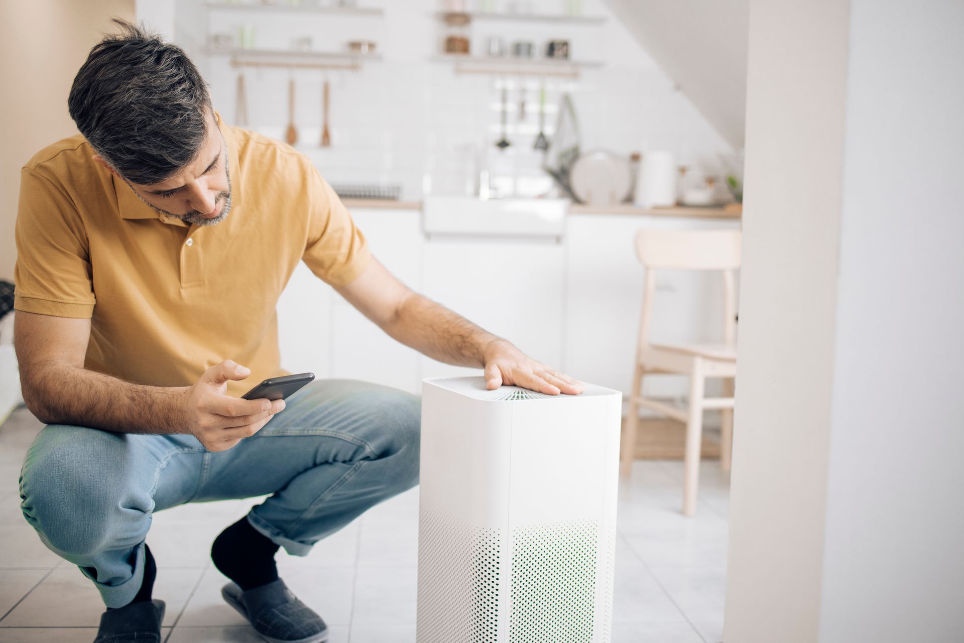 A man is kneeling next to a humidifier and looking at his phone.