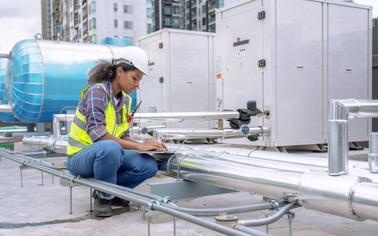 A woman is kneeling down on a roof looking at pipes and writing on a clipboard.
