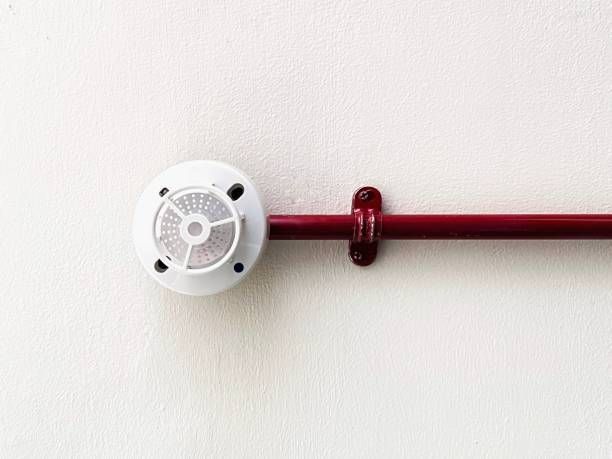 A fire alarm is attached to a red pipe on a white wall.