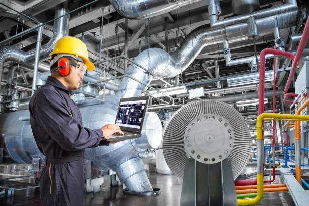 A man in a hard hat is using a laptop in a factory.