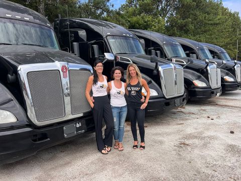 Three women are standing in front of a row of semi trucks.