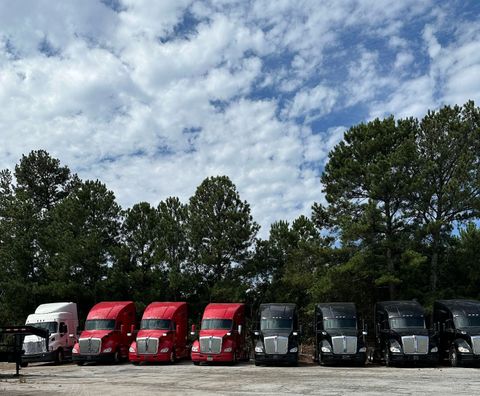 A row of semi trucks are parked in a lot with trees in the background.