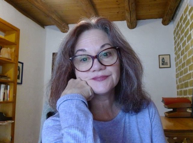 A woman wearing glasses is sitting in a room with her hand on her chin.